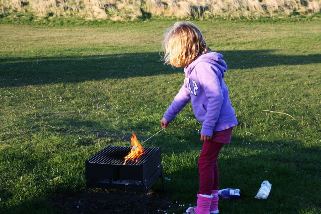 Young girl in a purple fleece is coking marshmallows in a fire made with sticks and branches