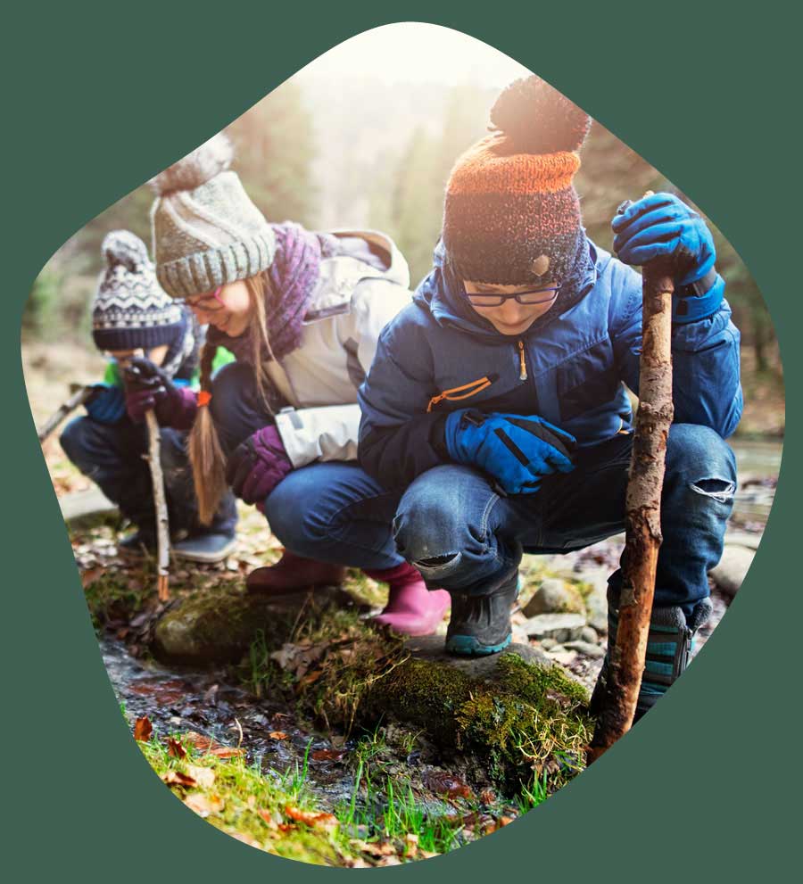 Three children are dressed in warm coats and hats as they play outdoors. They are exploring a burn and each holding sticks